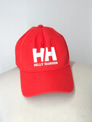 Röd keps från Helly Hansen - Snygg röd keps från Helly Hansen med böjd skärm och justerbar baksida. Kepsen har ett stort vitt HH-tryck och texten HELLY HANSEN framtill. Tillverkad i bomullstyg och har klassisk sexpanelsdesign med ventilationshål.
