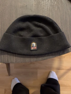 Parajumper Mössa - Säljer en svart ribbstickad mössa från Parajumper med uppvikt kant och en broderad patch framtill. Klassisk och enkel design som passar till det mesta. Perfekt för kalla dagar och enkel att matcha med olika outfits. Mössan är äkta.