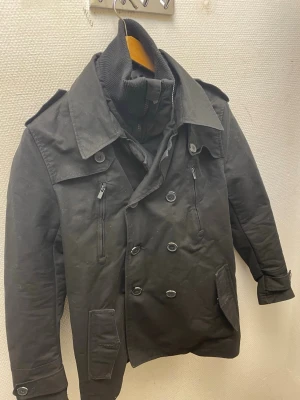 Svart skepparkavaj med hög krage - Stilren svart skepparkavaj med dubbelknäppning, bred krage och axelklaffar. Jackan har två bröstfickor med dragkedja, två fickor med lock och en hög ribbad innerkrage för extra värme. Klassisk modell som passar till många olika looks.