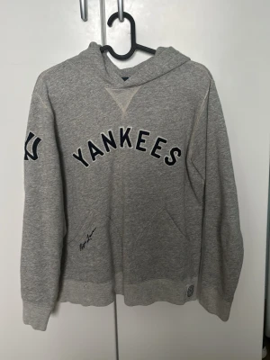 Grå Yankees hoodie Polo Ralph Lauren - Snygg grå hoodie från Polo Ralph Lauren med Yankees-tryck på bröstet och logga på ärmen. Tillverkad i mjuk bomullsblandning och har klassisk huva samt magficka. Del av Cooperstown Collection, perfekt för dig som gillar sportig stil.