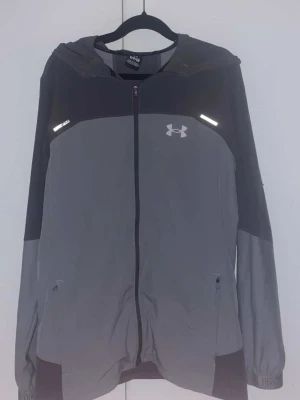 Grå vindjacka från Under Armour - Snygg grå och svart vindjacka från Under Armour med huva och hel dragkedja. Jackan har reflekterande detaljer, två fickor med dragkedja och logga på bröstet samt stor logga på ryggen. Tillverkad i lätt och funktionellt material, perfekt för aktiva dagar.