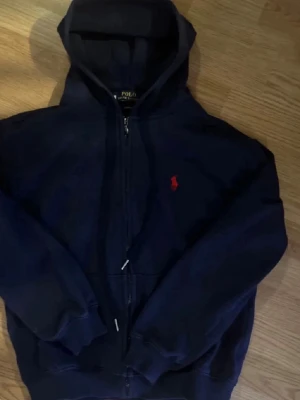  Polo Ralph Lauren hoodie - Mörkblå hoodie med dragkedja från Polo Ralph Lauren. Klassisk design med huva och fickor framtill. Ikoniska röda Polo-loggan broderad på bröstet. Tillverkad i mjukt bomullsmaterial som är skönt att bära.