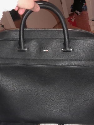 Svart datorväska från BOSS - Stilren svart datorväska från BOSS i strukturerat läder. Väskan har två robusta handtag, silverfärgade detaljer och en diskret logotyp framtill. Perfekt för att bära laptop och viktiga prylar till skolan eller jobbet.