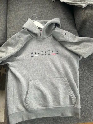 Grå Hilfiger hoodie med logga - Snygg grå hoodie från Tommy Hilfiger med stor logga och texten 'Hilfiger New York' på bröstet. Klassisk känguruficka framtill och huva. Tillverkad i mjukt sweatshirtmaterial, perfekt för en avslappnad och trendig look.