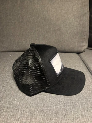 Svart truckerkeps med mesh - Säljer en svart truckerkeps med mesh på sidorna och bak, samt en böjd skärm. Framtill finns en vit patch utan synligt tryck. Kepsen har justerbar snapback och klassisk form. Perfekt för dig som gillar streetwear och enkel stil.
