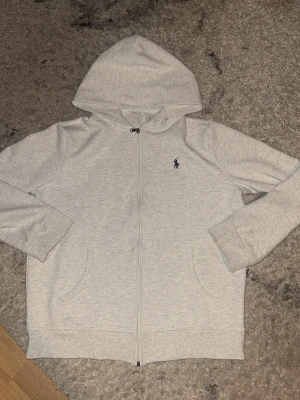 Ljusgrå hoodie från Polo Ralph Lauren - Snygg ljusgrå hoodie med dragkedja från Polo Ralph Lauren. Klassisk design med två fickor fram och mörkblå broderad logga på bröstet. Tillverkad i mjukt bomullsmaterial och har en stor huva för extra komfort.
