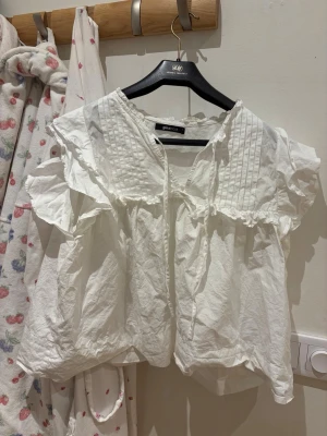 Vit blus med volanger från Gina Tricot - Söt vit blus från Gina Tricot med korta volangärmar och dekorativa veck framtill. Blusen har en lös passform, rundad halsringning med knytband och är tillverkad i ett lätt bomullstyg. Perfekt för en trendig och avslappnad look.