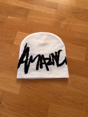 Vit mössa med svart Amarin-tryck - Säljer en vit mössa från Amarin med ett stort svart grafiskt tryck framtill. Mössan är mjuk och stretchig, perfekt för att hålla värmen och ge en cool streetwear-look. Passar dig som gillar enkel men ändå uttrycksfull stil.