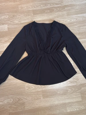Svart blus med spetsdetaljer - Svart blus med djup V-ringning och fina spetsdetaljer längs urringningen. Blusen är långärmad och har en märkerad midja. I storlek S/M
