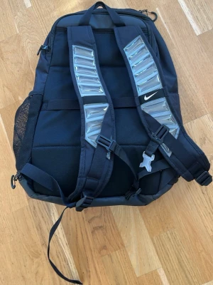 Svart Nike Elite ryggsäck - Snygg svart Nike Elite ryggsäck med stora vadderade axelband och ventilerande meshficka på sidan. Ryggsäcken har flera fack, dragkedjor och en tydlig Nike-logga samt ELITE-tryck på framsidan. Perfekt för träning, skola eller gymmet.