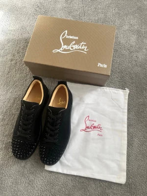 Svarta sneakers från Christian Louboutin - Snygga svarta sneakers från Christian Louboutin med klassiska röda sulor och svarta nitar framtill. Skorna har snörning och är tillverkade i skinn, vilket ger en lyxig känsla. Perfekta för dig som vill sticka ut med en exklusiv look. Diskutera gärna om priset, priset är ej hugget i stenen.