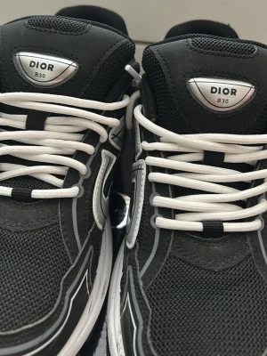 svarta sneakers - Snygga sneakers i svart mesh och mocka med vita och grå detaljer. Skorna har vita snören, reflekterande logga på sidan och en chunky vit sula. Perfekta för dig som vill ha en exklusiv och trendig look. Använda en gång och pris kan diskuteras. Tveka inte att höra av dig. Storlek 41 men passar 42