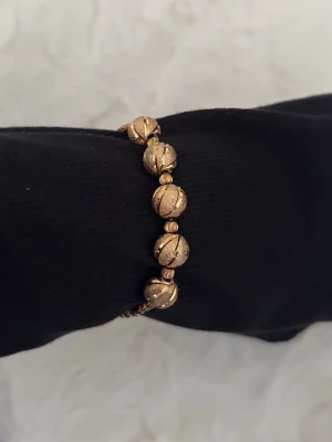 Guldigt armband med pärlor - Snyggt armband i guldfärg med fem större pärlor som har ett ingraverat mönster. Armbandet har en justerbar kedja och små guldfärgade detaljer mellan pärlorna. Perfekt accessoar för att lyfta din outfit. Köpt i London. Knappt använd. 