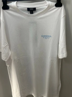 Vit t-shirt Sardinia SMOG - Vit t-shirt från SMOG med tryck på bröstet och en stor färgglad print på ryggen med motiv från Sardinien och texten 'La Dolce Vita'. Klassisk passform och rund halsringning. Perfekt för en chill och stilren look.
