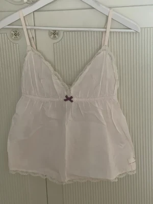 Ljusrosa linne från Odd Molly i storlek S i nyskick  - Söt ljusrosa linne med tunna axelband och spetsdetaljer längs kanterna från Odd Molly i storlek S i nyskick. Dekorerad med en liten lila rosett framtill. Tillverkad i ett tunt, mjukt tyg som känns luftigt och bekvämt. Justerbara axelband för bästa passform.