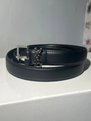 Svart skärp från Saint Laurent - Säljer ett svart skärp från Saint Laurent i skinn med silverfärgat YSL-logo och spänne. Skärpet har en klassisk och stilren design som passar till många outfits. Perfekt för dig som vill ha en exklusiv accessoar.