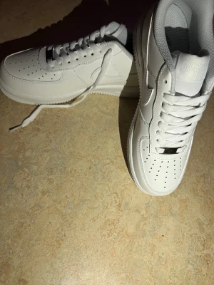 Nike Air Force 1 vita sneakers - Klassiska Nike Air Force 1 sneakers i helvitt läder med perforerad tå, vit sula och vita skosnören. Ikonisk design med Nike-logga på sidan och metallbricka på snörningen. Perfekta för en clean och stilren look.