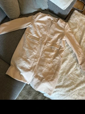 Ljusbeige stilren kappa med dragkedja - Snygg ljusbeige kappa med rak passform och dragkedja framtill. Kappan har två dekorativa ficklock och en enkel, minimalistisk design som passar perfekt till många outfits. Materialet ser ut att vara en mjuk och lätt tygblandning.
