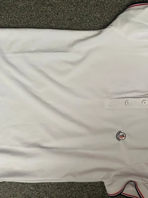 Vit pikétopp med rödblå detaljer - Säljer en vit pikétopp  med klassisk krage och knappar framtill. Toppen har-logga på bröstet och snygga rödblå ränder på krage och ärmslut. Perfekt för dig som gillar stilrena och exklusiva plagg.