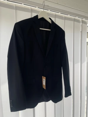 Svart Kavaj - Stilren svart kavaj med smal passform och enkla slag. Kavajen har två knappar framtill, tre knappar vid ärmslut och två fickor med lock samt en bröstficka. Perfekt för en dressad look. Aldrig någonsin använd, passar både kill eller tjej storlek 46/ mera XS 