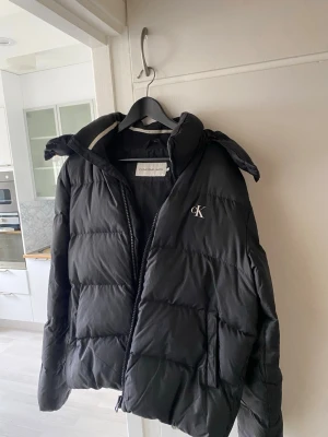 Svart pufferjacka från Calvin Klein - Svart pufferjacka från Calvin Klein med broderad CK-logga på bröstet. Jackan har hög krage, dragkedja framtill och två sidofickor. Perfekt för kalla vinterdagar och har en klassisk, stilren look. Materialet är glansigt och quiltat för extra värme.