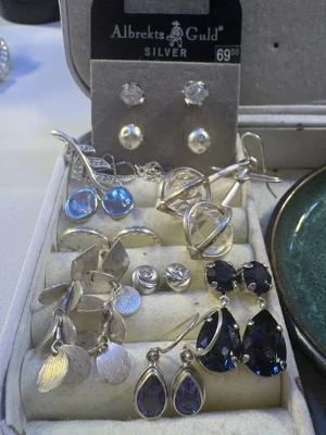 Mixade silvriga örhängen med stenar - Säljer ett set med flera par silvriga örhängen & halsband  från Albrekts Guld. Innehåller hängande modeller, vissa med blå, lila och klara stenar. Flera örhängen har droppformade eller runda detaljer och olika texturer. Perfekt för dig som gillar att variera din stil.