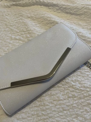 Vit glittrig kuvertväska med kedja - Snygg vit kuvertväska med diskret glittereffekt och silverfärgad metallkedja. Väskan har ett elegant metallspänne på framsidan och insidan är fodrad i matchande tyg. Perfekt för dig som vill ha en stilren accessoar med lite extra bling.