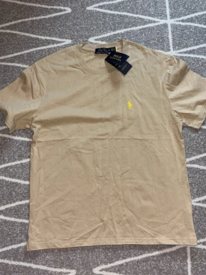 Beige t-shirt från Polo Ralph Lauren - Snygg beige t-shirt från Polo Ralph Lauren med klassisk gul broderad logga på bröstet. T-shirten har rund halsringning och korta ärmar, perfekt för en clean och stilren look. Tillverkad i mjuk bomull för maximal komfort.