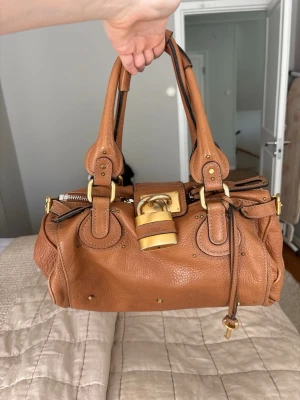 Chloé brun handväska i läder - Säljer en snygg brun handväska från Chloé i mjukt läder med guldfärgade metalldetaljer och ett stort dekorativt hänglås framtill. Väskan har dubbla handtag, dragkedja och flera spännande sömmar och nitar som ger en cool look.