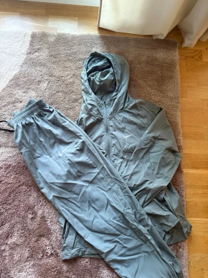 Grått Nike träningsset med huva - Säljer ett grått träningsset från Nike med matchande byxor och jacka. Jackan har huva och dragkedja framtill, byxorna har resår i midjan och smal passform. Perfekt för träning eller chill. Materialet är lätt och andas bra.
