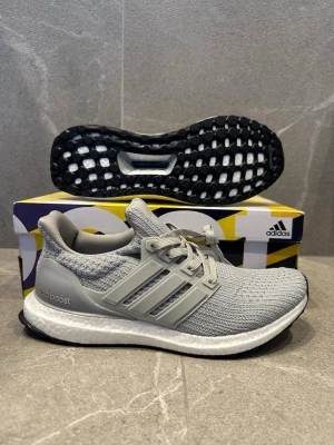 Adidas Ultra Boost grå sneakers - Grå Adidas Ultra Boost sneakers med stickad ovandel och klassiska tre ränder i plast på sidorna. Skon har en vit Boost-mellansula och svart yttersula. Perfekt för dig som gillar sportig och stilren look. Skön passform med snörning och mjuk känsla.