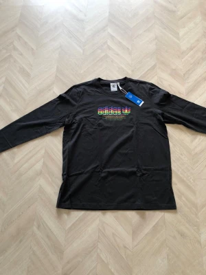 Svart långärmad t-shirt från Adidas - Svart långärmad t-shirt från Adidas med färgglatt tryck och logga på bröstet. Klassisk rund halsringning och bekväm passform. Tillverkad i mjuk bomull, perfekt för en avslappnad och sportig stil.