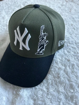 Olivgrön New Era Yankees keps - Snygg olivgrön keps från New Era med New York Yankees-logga och Frihetsgudinnan broderad framtill. Svart böjd skärm och justerbar baksida. Broderad text 'New York' bak. Perfekt för dig som gillar streetstyle och sportiga accessoarer.