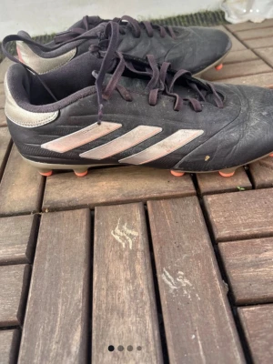 Svarta Adidas fotbollsskor med dobbar - Säljer ett par svarta fotbollsskor från Adidas med klassiska tre vita ränder på sidan. Skorna har snörning, orange detaljer på sulan och insidan samt dobbar för spel på gräs. Ovandelen är i syntetmaterial och designen är smidig och sportig.