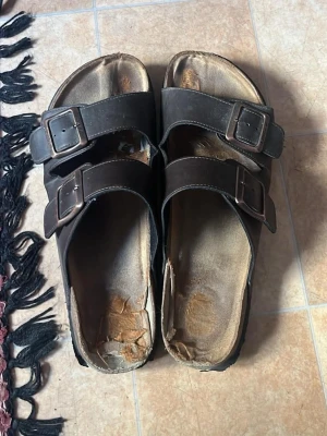Bruna sandaler med dubbla spännen - Slitna ”slit och släng” sandaler. Bruna sandaler med två breda remmar och justerbara metallspännen. Klassisk slip-in modell med öppen tå och fotbädd. Perfekta för en avslappnad stil och passar till många outfits. Tror att storleken är 43, inte helt säker.