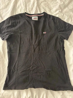 Svart t-shirt från Tommy Jeans - Svart t-shirt från Tommy Jeans med klassisk rund hals och liten broderad logga på bröstet. T-shirten har en enkel och stilren design med diskreta detaljer i rött, vitt och blått på insidan av kragen. Tillverkad i mjuk bomull.