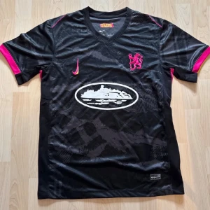 Corteiz Chelsea tröja - Snygg svart sporttröja med rosa och gula detaljer, V-ringning och korta ärmar. Tryckt emblem på bröstet och ett stort vitt tryck på magen. Perfekt för träning eller match. Materialet är lätt och andas.