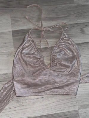 Glittrig champagnefärgad halterneck topp - Supersnygg glittrig champagnefärgad halterneck topp från Gina Tricot. Toppen har knyt i nacken och omlott framtill med en croppad passform. Perfekt för dig som vill sticka ut lite extra med metallic vibe.