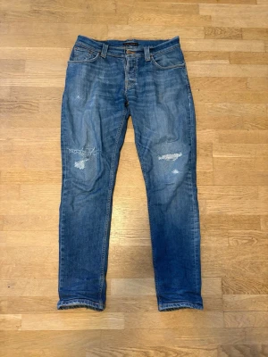 Blå slitna jeans från Nudie Jeans - Säljer ett par blå jeans från Nudie Jeans med snygga slitningar och lagade detaljer på knäna. Klassisk femficksmodell med raka ben och normal midja. Perfekta för en avslappnad och trendig look. Archive L:108cm