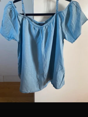 Ljusblå offshoulder topp från Lindex - Snygg ljusblå offshoulder topp från Lindex med korta ärmar och tunna axelband. Toppen har en avslappnad passform och är tillverkad i mjuk bomull, perfekt för varma dagar.