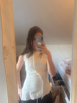 Vit ärmlös topp från Weekday - Snygg vit/beige ärmlös topp från Weekday med asymmetrisk design och råa kanter. Toppen har en lätt rynkad struktur och figurnära passform, perfekt för att lyfta din outfit med en modern touch.