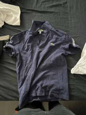 Marinblå pikétröja från Lacoste - Klassisk marinblå pikétröja från Lacoste med korta ärmar och den ikoniska krokodilloggan på bröstet. Tröjan har krage och knappar vid halsen. Perfekt för en avslappnad men ändå stilren look.