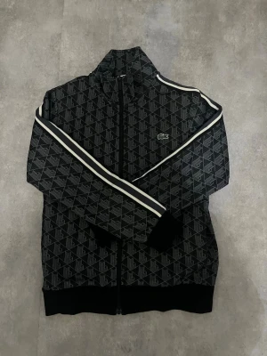 Svart mönstrad ziptröja från Lacoste - Snygg svart ziptröja från Lacoste med grått geometriskt mönster och vita ränder längs ärmarna. Tröjan har hög krage, dragkedja framtill och den klassiska Lacoste-loggan på bröstet. Perfekt för en sportig och trendig look.