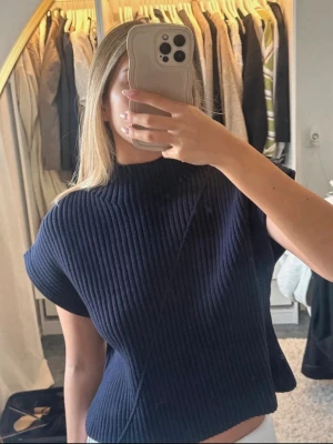 Mörkblå ribbad topp med kort ärm - Säljer en mörkblå ribbad topp med korta ärmar och hög hals. Toppen har en loose passform och är tillverkad i ett stickat material som ger en snygg struktur. Perfekt att styla med jeans eller kjol för en trendig look.