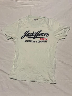 Vit Jack & Jones t-shirt med tryck - Snygg vit t-shirt från Jack & Jones med stort tryck framtill i marinblått och rött där det står 'Jack & Jones Denim Clothing Company'. Klassisk passform och rund halsringning. Perfekt till jeans eller shorts. Bild när den sitter på finns vid önskan! 