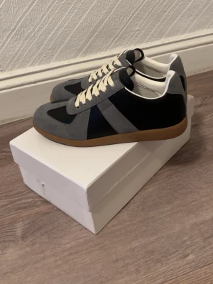 Maison margiella skor - Snygga sneakers i grått och svart med ovandel i mocka och textil, vita skosnören och klassisk gummisula. Perfekt för dig som gillar stilrena och tidlösa skor med en modern twist.
