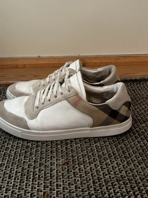 Burberry vita låga sneakers - Snygga vita sneakers från Burberry med klassiskt rutigt mönster på sidan. Skorna har detaljer i mocka och läder, samt vita snören och sula. Perfekta för dig som vill ha en stilren och tidlös look med en touch av lyx.