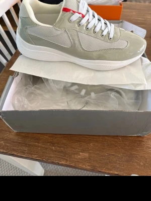 Beige sneakers från Prada - Snygga beige sneakers från Prada med vita detaljer och vit sula. Skorna har en mix av mesh och glansigt material, samt röda och vita accenter vid plösen. Klassisk låg modell med vita skosnören och diskret logga. Lite defekt på framsidan av en av skorna se bilder.