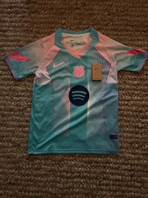 Nike FC Barcelona matchtröja turkos - Säljer en FC Barcelona matchtröja från Nike i turkos, ljusblå och rosa färger. Tröjan har V-ringning, klubbmärke och Nike-logga på bröstet samt Spotify-logga i mitten. Tillverkad i lätt och ventilerande funktionsmaterial, perfekt för fotboll eller träning.