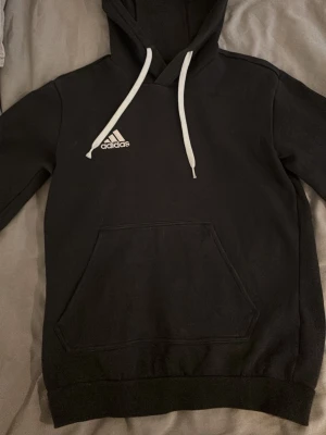 Svart hoodie från Adidas med huva - Svart hoodie från Adidas med vit logga på bröstet och vita dragsnören i huvan. Klassisk känguruficka framtill och ribbade muddar vid ärmslut och nederkant. Tillverkad i mjukt material som passar perfekt för en avslappnad stil.